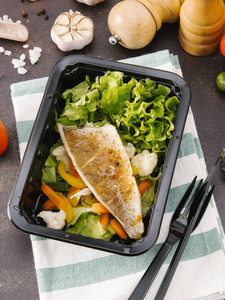 烤鱼片配菜沙拉在黑盘子里用叉子和刀在条纹餐巾上