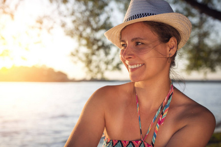 微笑愉快美丽的晒黑夫人在夏天帽子在湖岸边反对日落的背景在地平线。可爱的海滩女孩