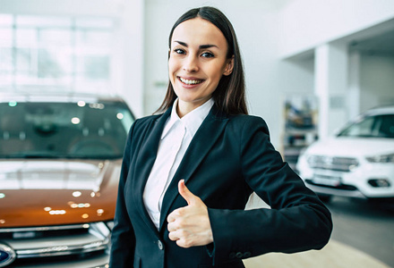 黑色西装的开朗微笑的商人的肖像显示拇指站在经销商汽车背景