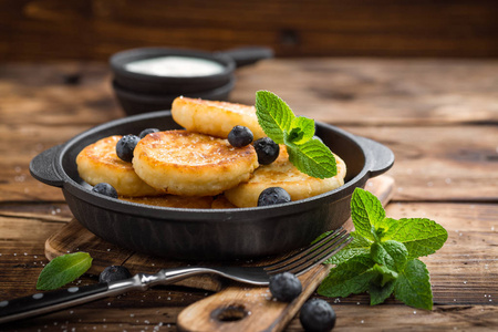 美味的奶酪薄饼或 syrniki 与新鲜的蓝莓在铸铁平底锅上深色木制仿古背景。可口的早餐