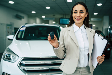 漂亮的微笑的女售货员与汽车钥匙和文件在手上的汽车经销商的背景