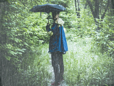 人在大衣用雨伞和袋子徒步旅行在森林的山在路