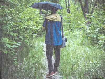 人在大衣用雨伞和袋子徒步旅行在森林的山在路