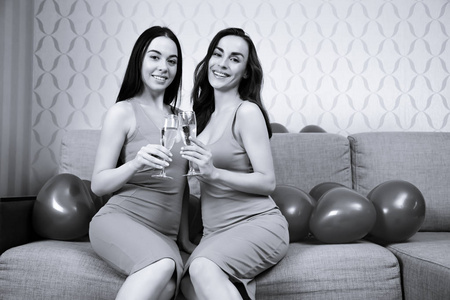 母鸡党。两个美丽开朗的女孩朋友在明亮的晚礼服喝香槟和休息在一个聚会上坐在沙发上