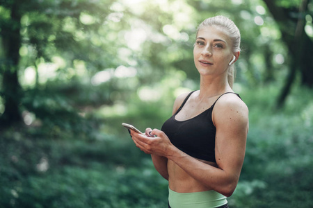 妇女赛跑者在夏天早晨公园听音乐在智能手机使用蓝牙耳机。女性健身女孩在外面的小路上慢跑