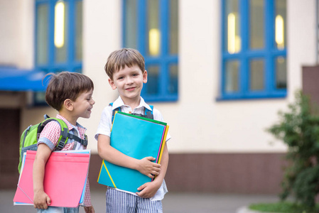 可爱小学校学生轻快地谈校园。孩子们有个好心情。温暖的春天的早晨。后面在同学书包的肩。在手中的男孩明亮的红色文件夹之一