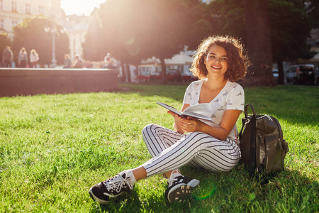美丽的大学女生在校园公园看书。快乐女学生学习户外坐在草地上