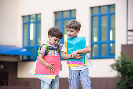 可爱小学校学生轻快地谈校园。孩子们有个好心情。温暖的春天的早晨。后面在同学书包的肩。在手中的男孩明亮的红色文件夹之一