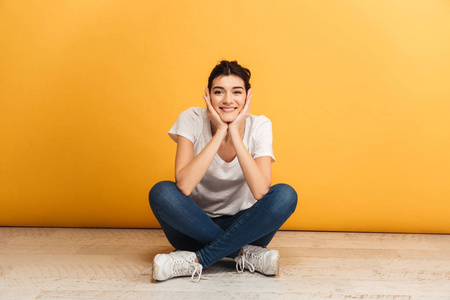 一个快乐的年轻女子的肖像坐在地板上的腿交叉, 看着在黄色背景相机
