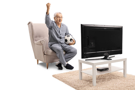 快乐的成熟男子穿着睡衣坐在扶手椅上看足球电视和握住他的手在白色背景上孤立