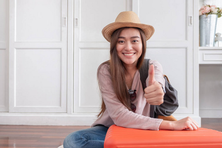年轻的亚洲女性旅行者竖起大拇指带着橙色的手提箱准备回家度假. 背包客旅游理念