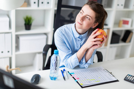 一个年轻的女孩坐在办公室的桌子上, 手里拿着一个苹果。