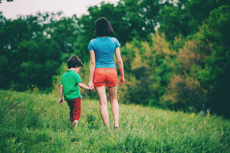 男孩和他的母亲在草地上散步。一个女人牵着她儿子的手。孩子和妈妈正沿着田野走。绿草