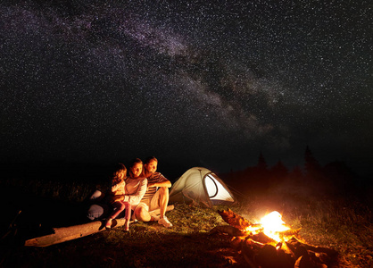 晚上在山上露营。旅游家庭妈妈, 爸爸和女儿坐在一个日志附近的照明帐篷和燃烧篝火下惊人的星空和银河。旅游业和旅游概念