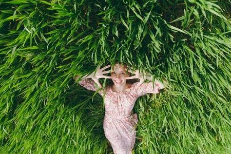 年轻的微笑美丽的女人穿着浅色图案的衣服躺在草地上显示双手仰望在阳光明媚的天气在田野上明亮的绿色背景。春天自然。生活方式, 休闲概