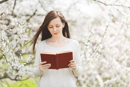 年轻的微笑放松温柔的女人在轻休闲的衣服阅读有趣的书站在城市花园或公园在盛开的树木背景。春天的自然, 花朵。生活方式, 休闲概念