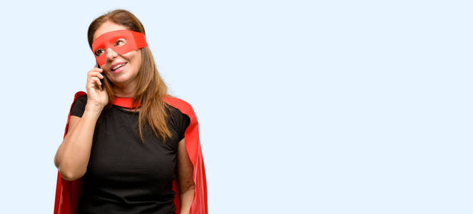 中年超级英雄女子戴着红面具和海角快乐的谈话使用智能手机孤立的蓝色背景