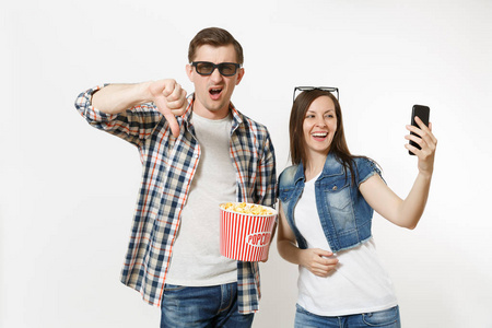 年轻夫妇, 妇女和男子在3d 眼镜看电影的日期持有的爆米花桶自拍在手机上显示拇指向下孤立的白色背景。情感在戏院