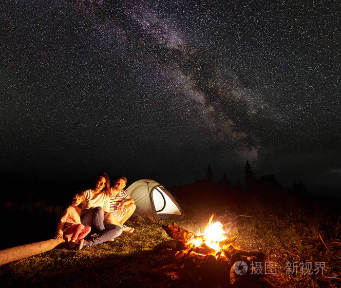 家庭露营在山上的夜晚, 坐在一个日志前的照明帐篷和燃烧篝火下美丽的