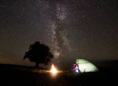 在山上露营的夜晚。年轻的旅游妇女坐在门口的照明帐篷, 有休息附近的篝火在美丽的深星空和银河。旅游和旅游概念