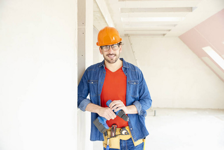 年轻修理工戴安全帽和工具带的肖像在工地施工时使用锤钻驱动程序