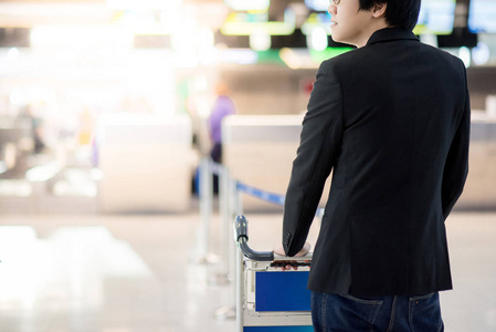 年轻的亚洲商人与他的行李在机场手推车等待登机柜台在国际机场航站楼, 商务旅行的概念
