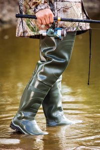 渔夫站在橡胶靴里, 为河里的沼泽, 夏天手里拿着一个鱼竿。