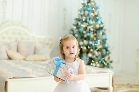 小可爱女孩保持礼物, 穿白色礼服和站立在卧室与圣诞树