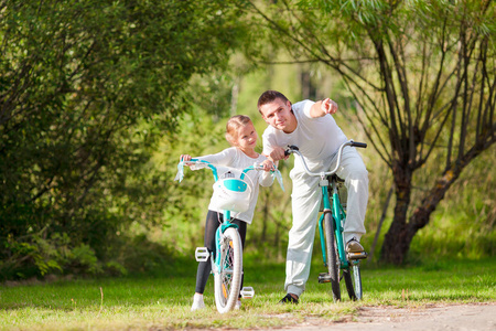 年轻的父亲和小女孩骑自行车在夏季温暖的一天。年轻活跃家庭骑自行车