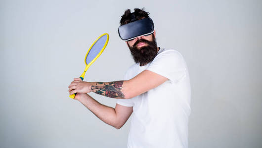 戴着虚拟眼镜的家伙打网球和球拍和球。有胡子的男人在 Vr 眼镜打网球, 灰色背景。时髦在繁忙的面孔使用现代技术为体育游戏。虚拟体
