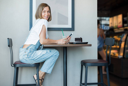 年轻的微笑的女人穿着工作服白色 t恤。坐餐桌咖啡厅吃咖啡蛋糕。使用手机。灰色墙壁