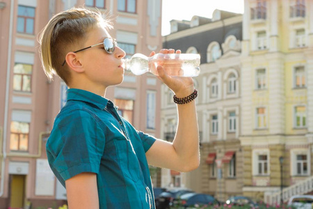 少年男孩喝水从瓶子在晴朗的炎热的夏天在城市