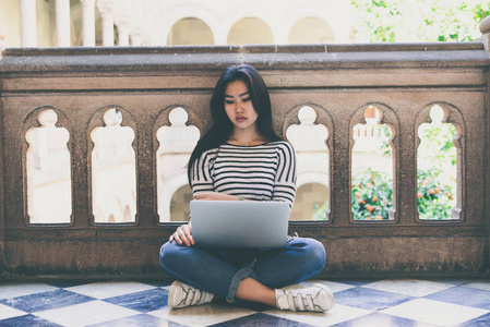 亚洲学生女孩穿着休闲装, 坐在大学校园阳台的地板上, 在笔记本电脑上工作