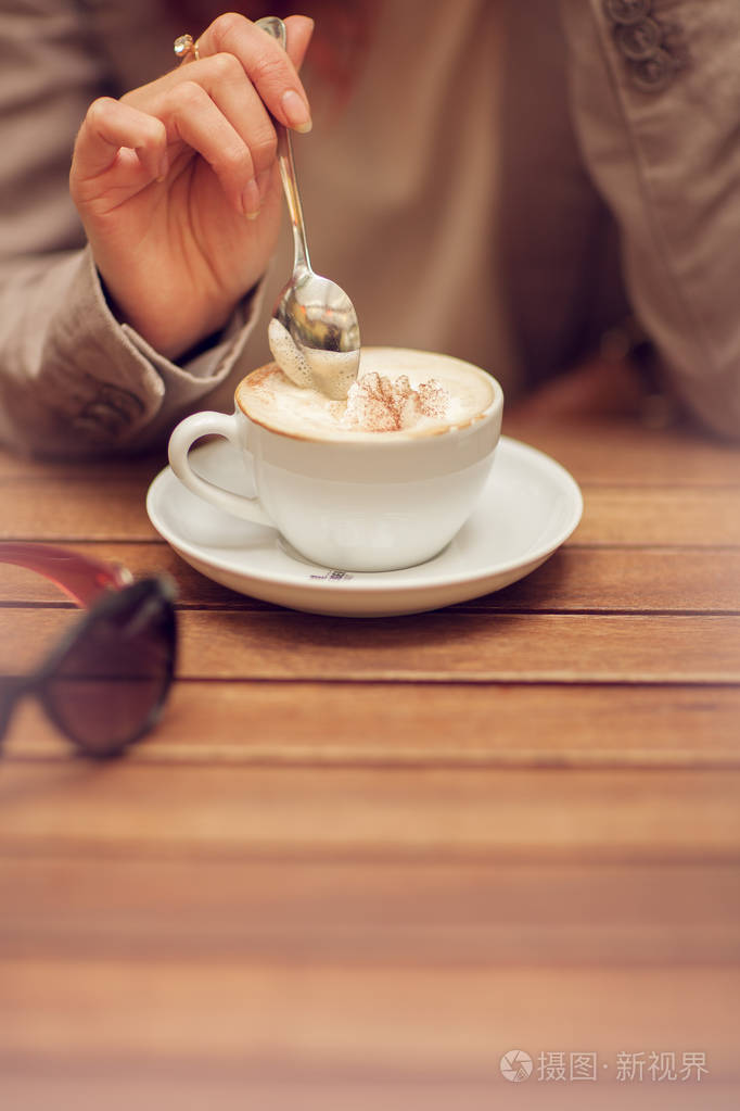 年轻女子手中拿着勺子, 在一张木桌上的咖啡厅里搅动一杯咖啡, 可以