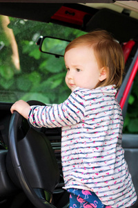 孩子在汽车方向盘上玩耍。可爱的小女孩驾驶汽车。孩子站在大汽车。关闭年轻的小女婴转方向盘的汽车。儿童造型与肖像概念