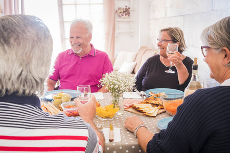 一群老年人在厨房吃奶酪, 胡萝卜和墨西哥食物, 一起欢笑, 玩得开心。为聚会和愉快的时间喝葡萄酒. 窗背光源