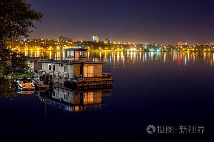小船驻地与房子在水在湖或河在晚上, 反射夜城市光在水中