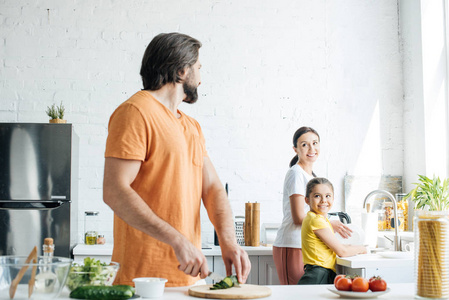 有吸引力的年轻父亲切黄瓜沙拉, 而他的妻子和女儿在厨房洗盘子