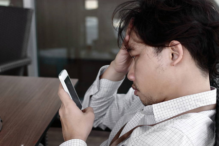 沮丧疲惫的亚洲商人用手在额头上寻找移动智能手机在他的手在办公室
