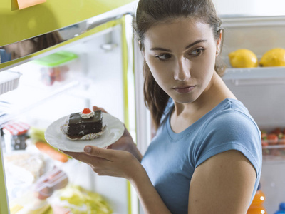 饥饿的年轻女子从冰箱里拿出美味的甜点, 吃点心, 节食失败的概念