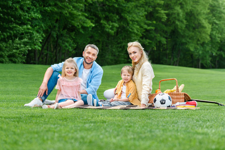 愉快的家庭与二个孩子微笑在照相机, 而坐在一起在公园野餐