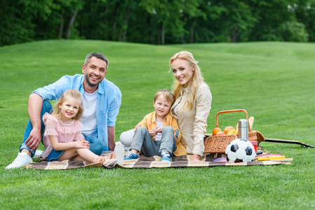 愉快的家庭与二个孩子微笑在照相机, 而坐在一起在格子在公园野餐