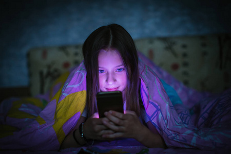 小女孩在床上在晚上的毯子下看着智能手机