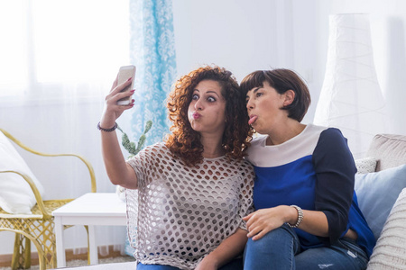 两名年轻女性在家里用手机拍照, 做着漂亮有趣的表情。爱与朋友在网上交流的最好朋友