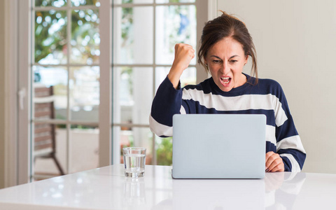 中年妇女用膝上型电脑在家里恼怒和沮丧的呼喊与愤怒, 疯狂和叫喊与举手, 愤怒的概念