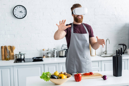 微笑的胡子年轻人在围裙使用虚拟现实耳机, 而在厨房做饭