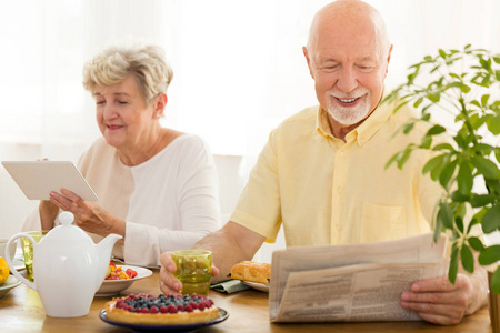 快乐的老年妇女使用片剂和她的丈夫读报纸