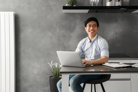 一个开朗的年轻亚裔男子的肖像坐在桌子上的笔记本电脑在家里的文书工作