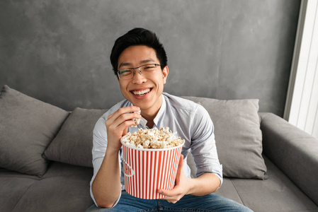 一个开朗的年轻亚裔男子的肖像吃爆米花, 而坐在沙发上, 在家里看电视