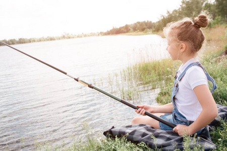 一张女孩独自坐在河岸边的照片。她在钓鱼。女孩用双手握住鱼杆。她正在看水。女孩是非常严肃的